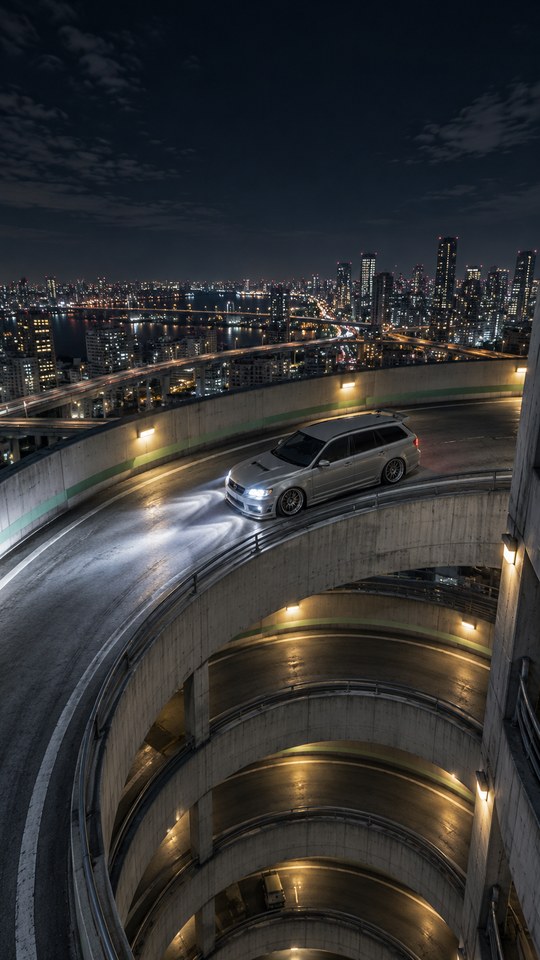 A pearl-grey unbranded tuner wagon climbs a spiral parking ramp at night, viewed from high above through the open central void.